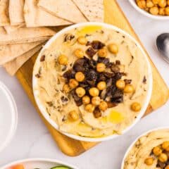 A bowl of hummus topped with roasted chickpeas and caramelized onions, surrounded by pita bread slices, cucumber rounds, carrot sticks, and a small bowl of extra chickpeas on a white surface.