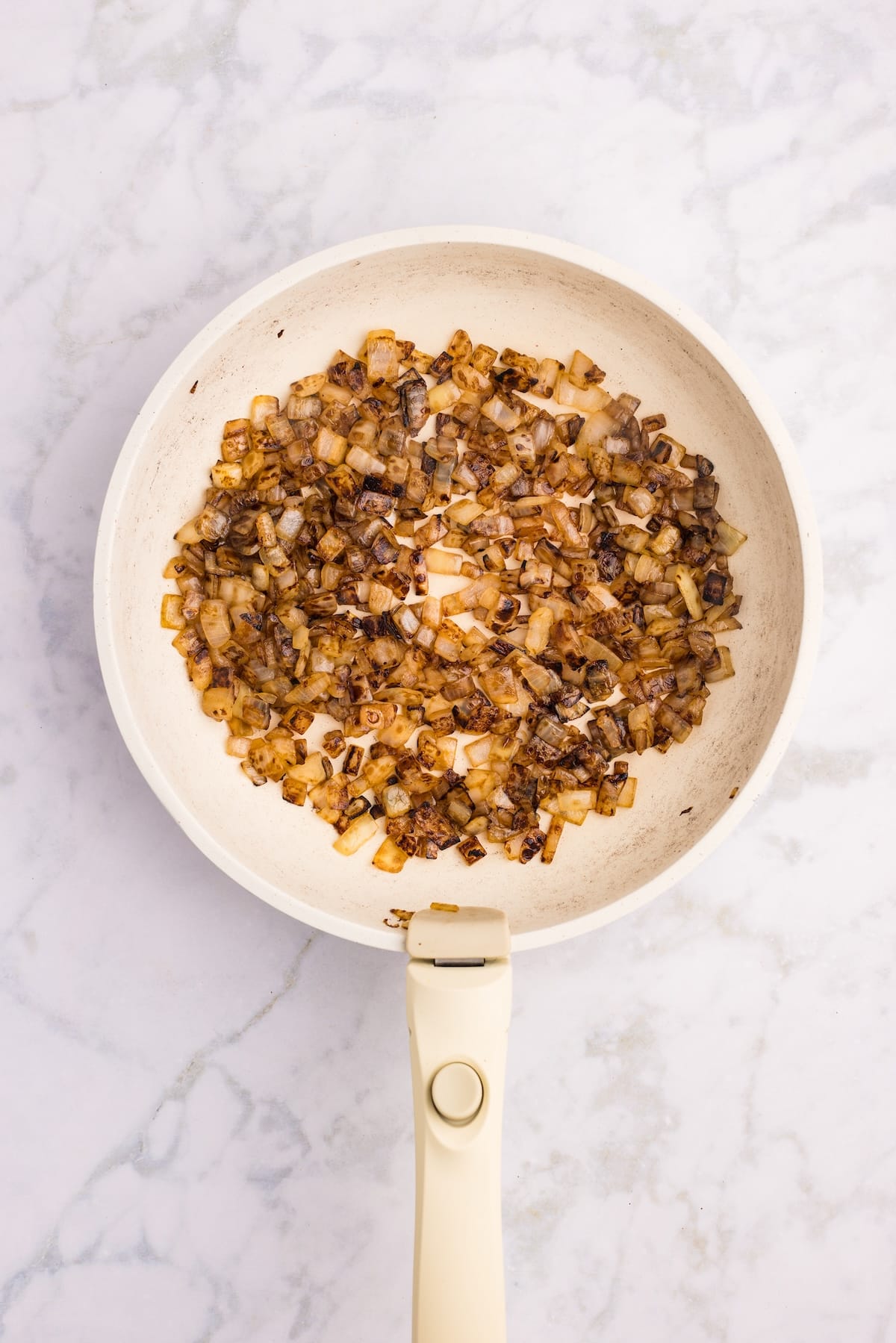 A white frying pan with browned, sautéed onions sits on a light marble surface. The onions are evenly cooked and golden with some darker caramelized pieces.