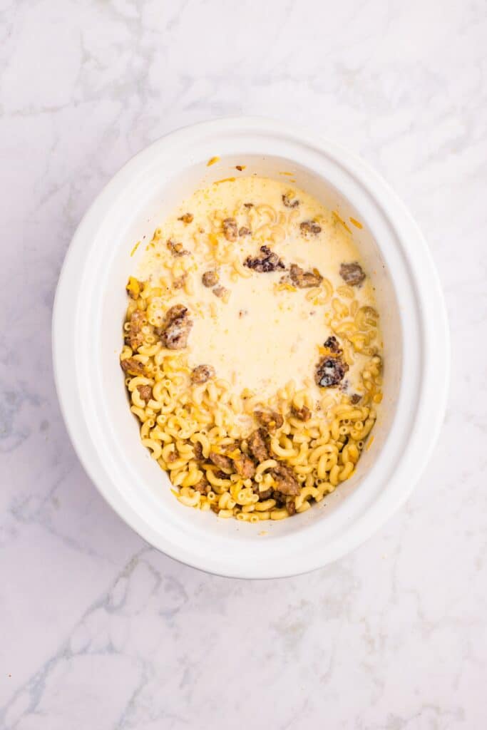 A white slow cooker filled with uncooked macaroni pasta, ground beef, and a creamy sauce mixture, viewed from above on a light marble surface.