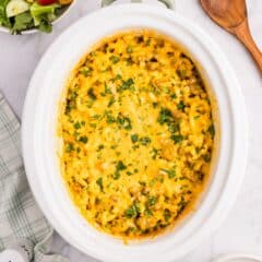 A creamy macaroni and cheese dish in a slow cooker, topped with chopped parsley. Surrounding it are a green-striped towel, salt and pepper shakers, a salad bowl, shredded cheese, and a wooden spoon.