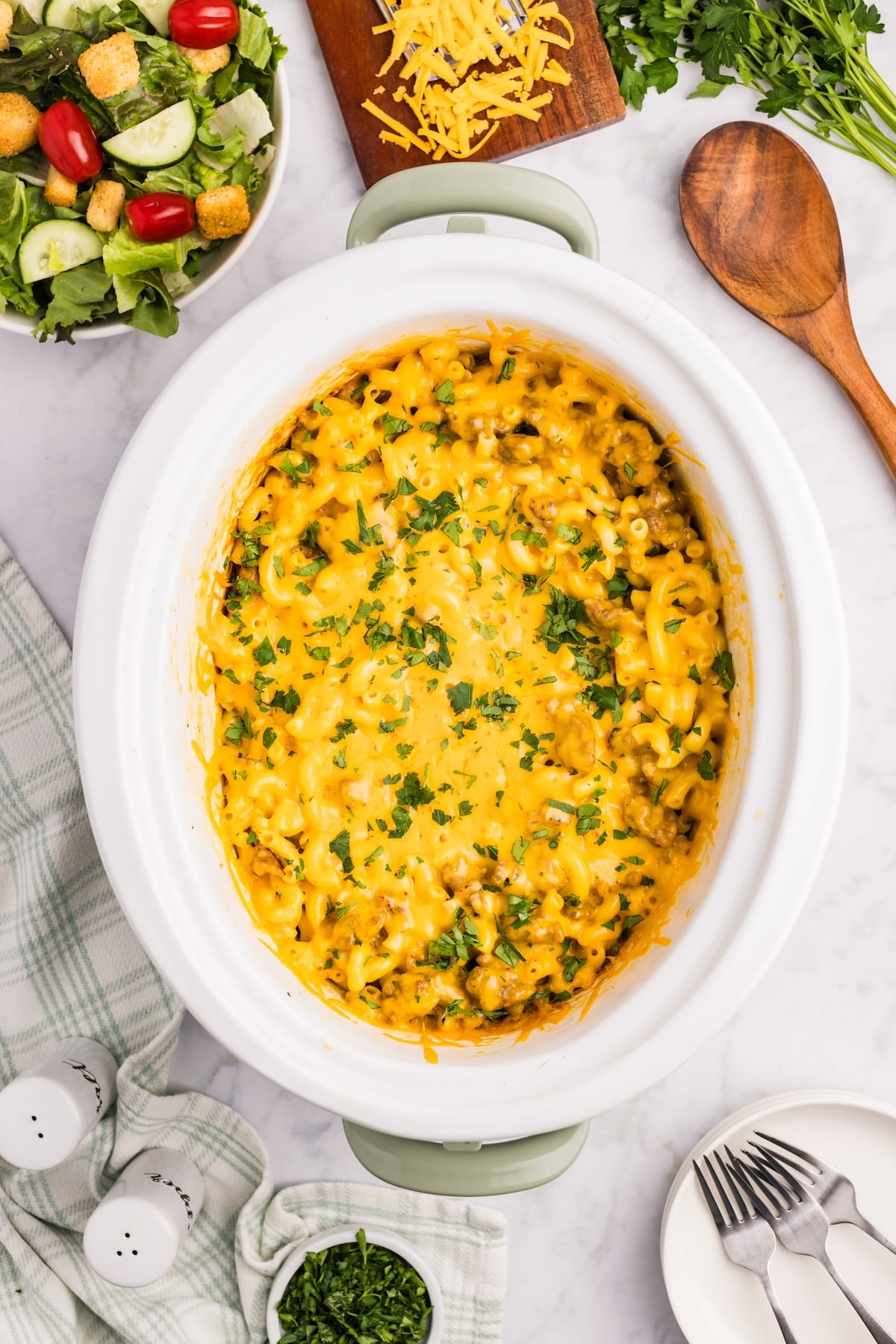 A creamy macaroni and cheese dish in a slow cooker, topped with chopped parsley. Surrounding it are a green-striped towel, salt and pepper shakers, a salad bowl, shredded cheese, and a wooden spoon.