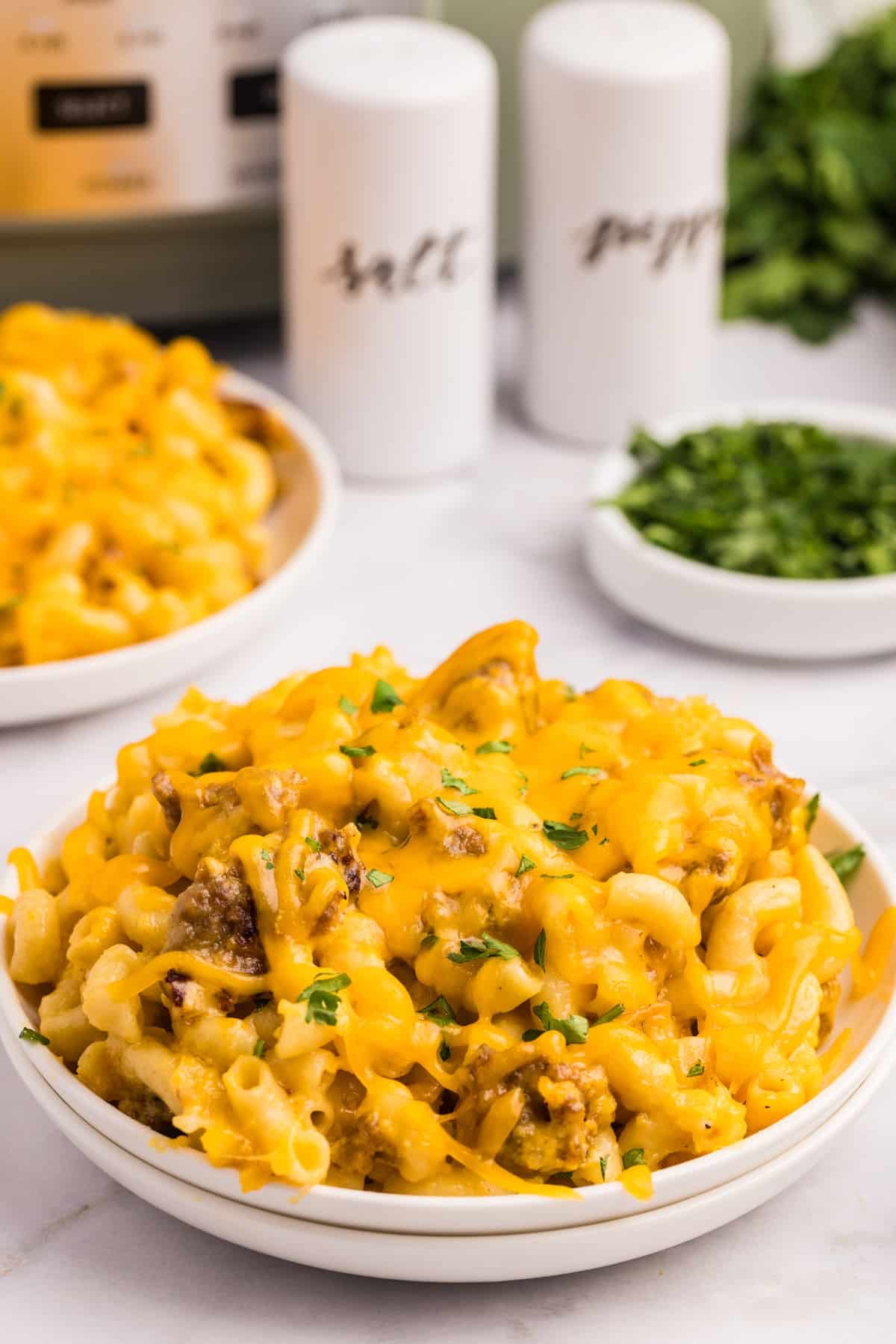 A white plate filled with cheesy macaroni and ground beef casserole, topped with melted cheddar and chopped herbs. In the background are salt and pepper shakers, a bowl of chopped parsley, and another plate of casserole.