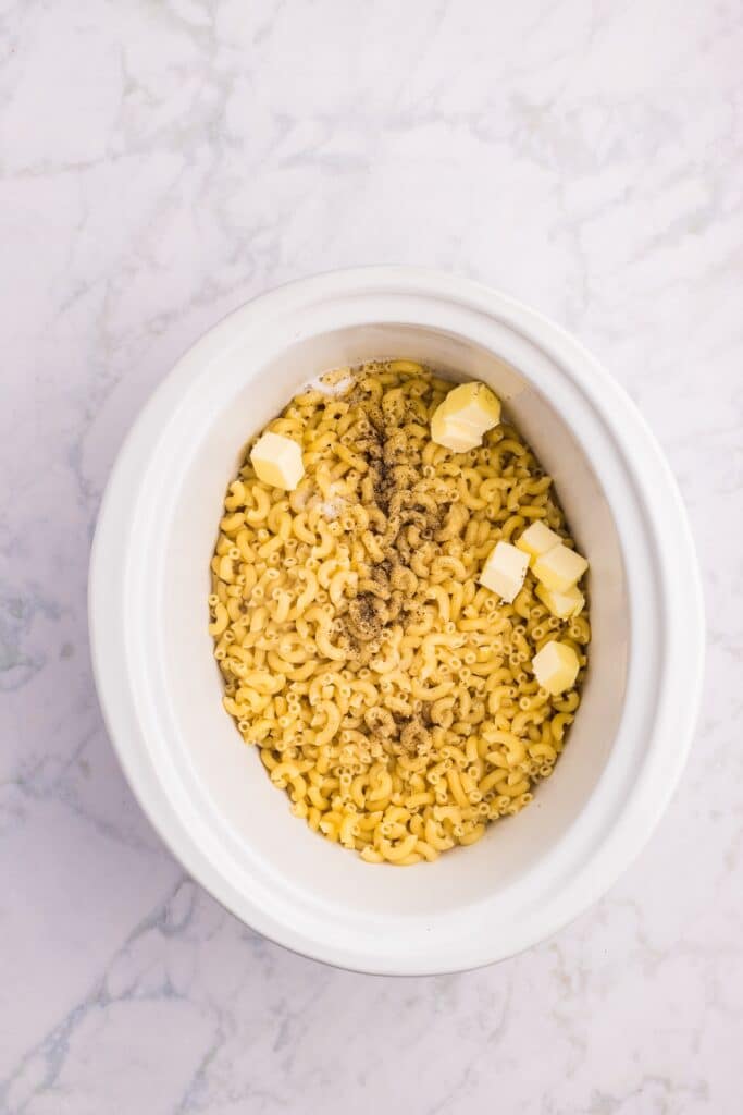 Uncooked elbow macaroni, cubes of butter, and black pepper in a white oval slow cooker on a light marble surface.
