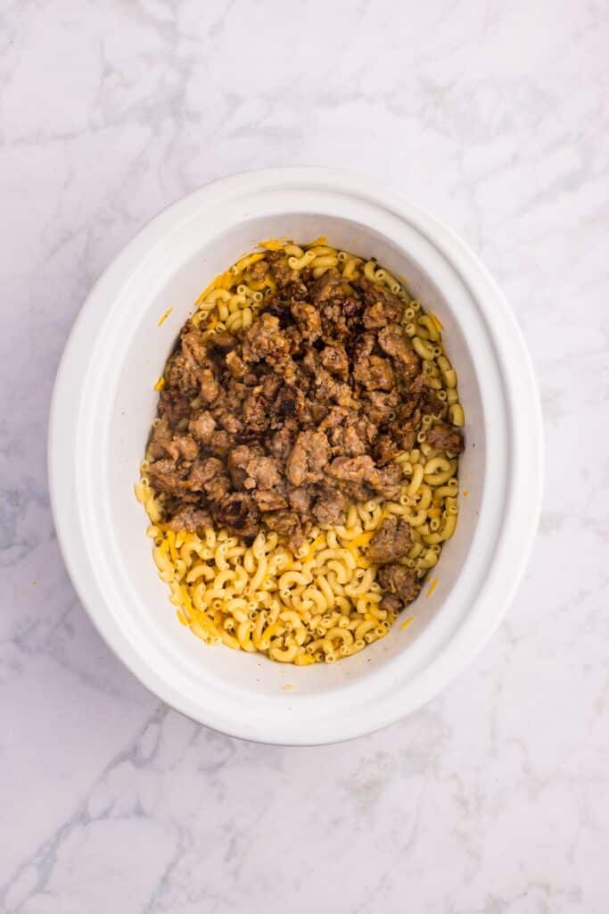A white slow cooker filled with cooked macaroni pasta and browned ground beef, placed on a light-colored marble surface.