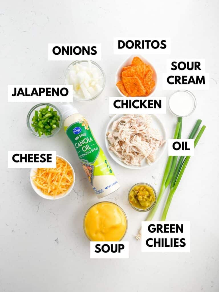 Ingredients for a recipe are laid out on a white surface, labeled: diced onions, Doritos chips, sour cream, shredded chicken, oil spray, green onions, canned green chilies, condensed soup, shredded cheese, and chopped jalapeno.