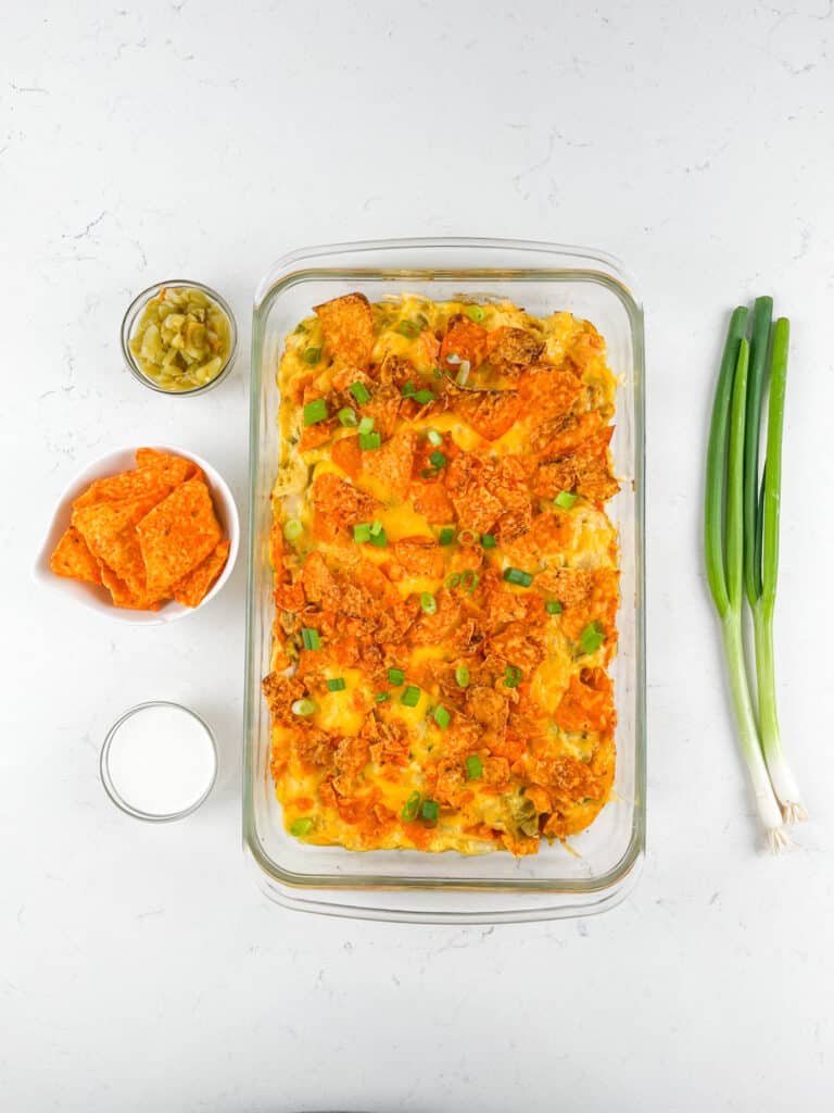 A glass baking dish filled with a cheesy casserole topped with crushed orange chips and green onions, next to a bowl of orange chips, a bowl of chopped pickles, a glass of milk, and three green onions on a white surface.