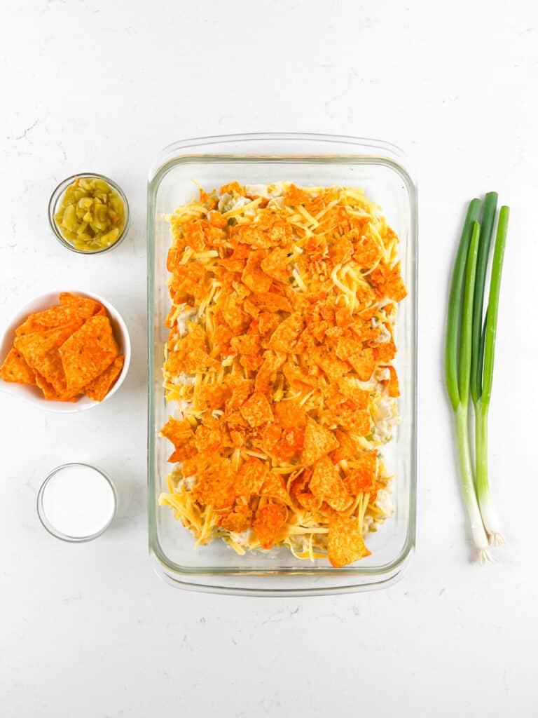 A glass baking dish with a casserole topped with crushed orange chips sits on a white surface. Surrounding it are bowls of sliced jalapeños, orange chips, a glass of milk, and three fresh green onions.