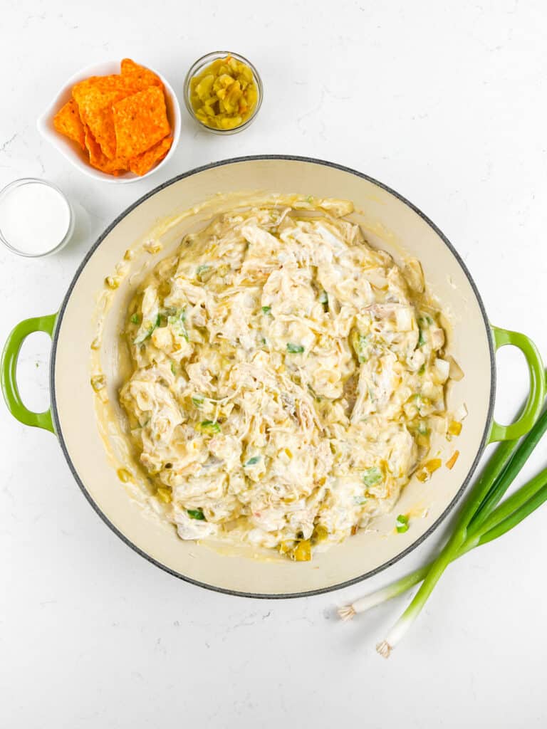 A green pot filled with creamy shredded chicken mixture, surrounded by small bowls of orange chips, chopped green peppers, a cup of milk, and green onions on a white surface.