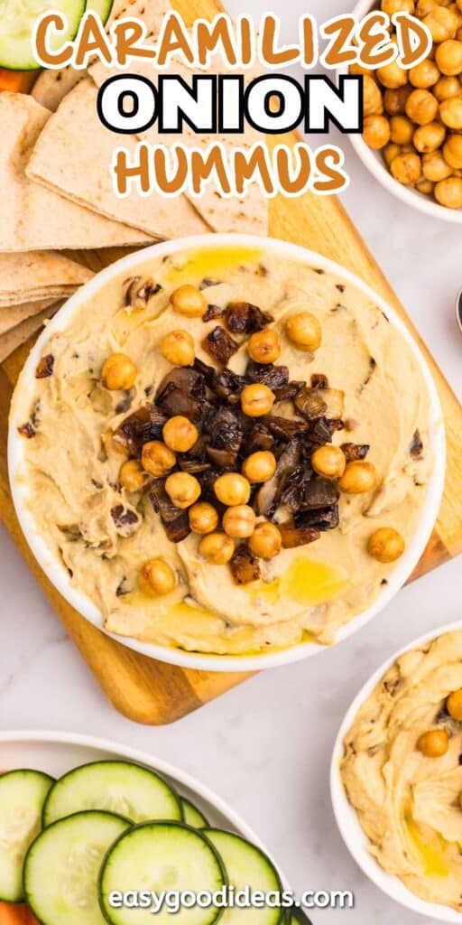 A bowl of hummus topped with caramelized onions and chickpeas sits on a wooden board, surrounded by pita bread slices and cucumber rounds. Text reads Caramelized Onion Hummus and easygoodideas.com.