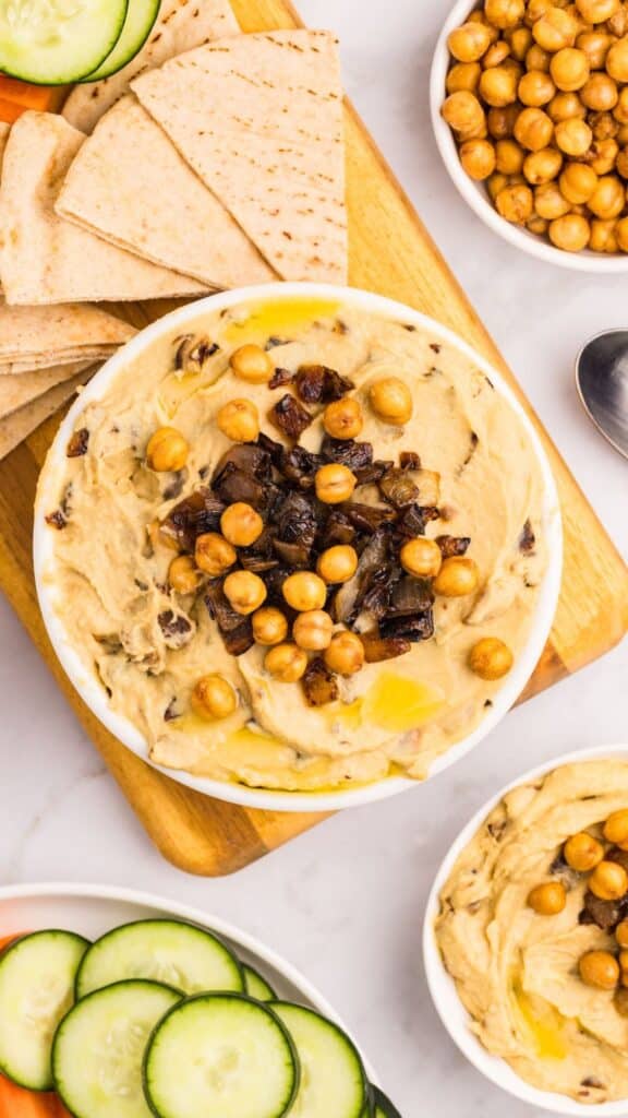 A bowl of hummus topped with chickpeas and chopped dates, drizzled with olive oil, sits on a wooden board with pita bread, cucumber slices, and a spoon nearby.
