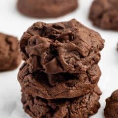 A stack of three thick, chocolate cookies with visible chocolate chunks sits on a white surface, with more cookies blurred in the background.