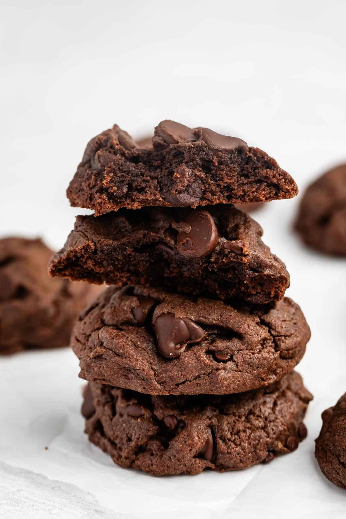 A stack of three thick, chocolate chocolate-chip cookies on white parchment paper, with the top cookie broken in half to reveal a soft, gooey center. More cookies are blurred in the background.