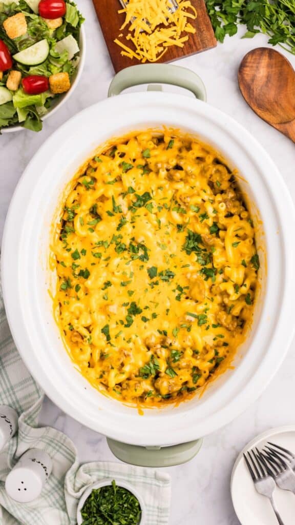 A crockpot filled with cheesy macaroni and ground beef, garnished with chopped parsley. Surrounding the crockpot are salad, shredded cheese, a wooden spoon, and utensils on a marble counter.