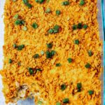 A glass baking dish filled with a cheesy casserole topped with crushed orange chips and chopped green onions, with one serving missing from the corner.