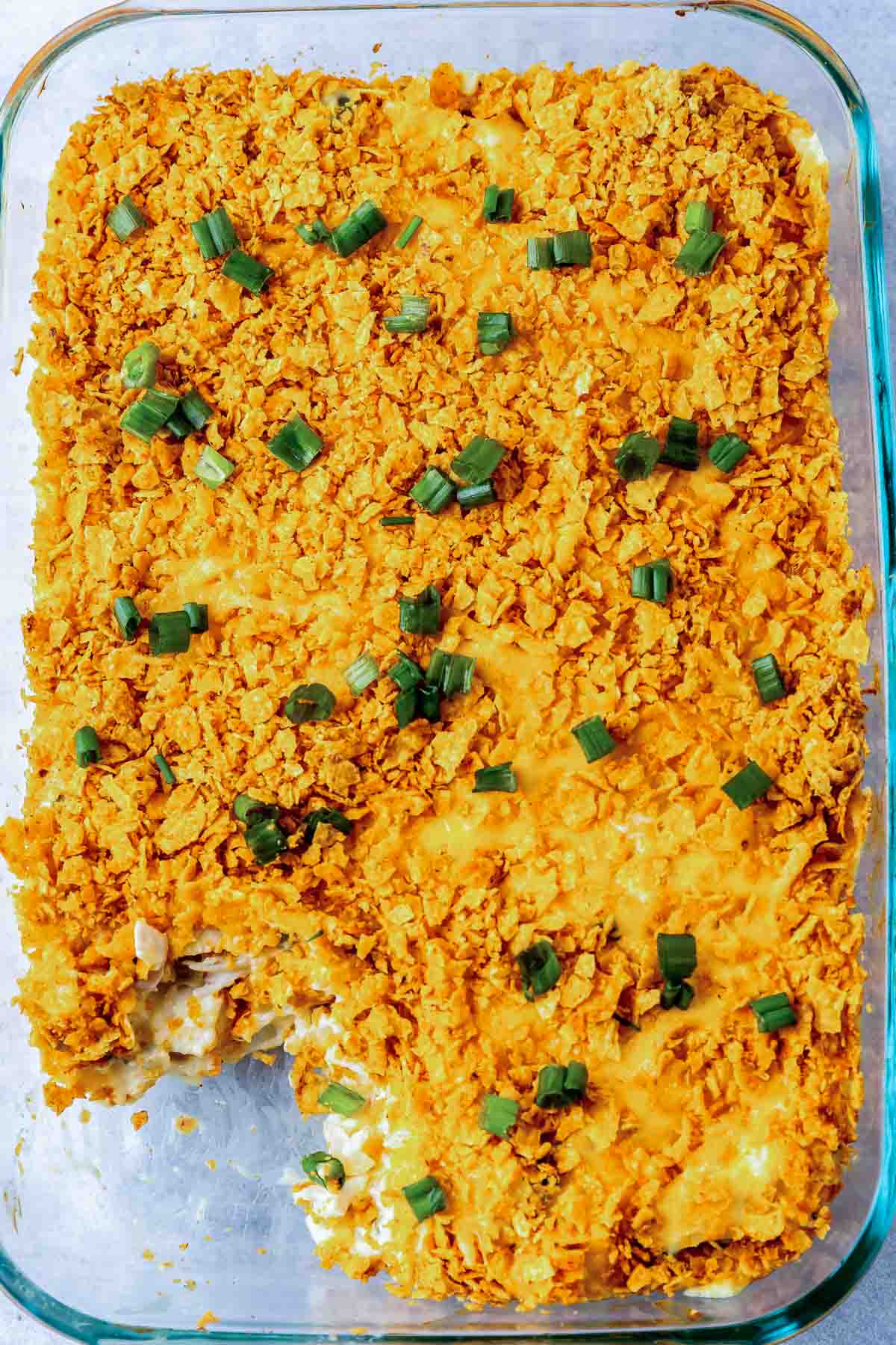 A glass baking dish filled with a cheesy casserole topped with crushed orange chips and chopped green onions, with one serving missing from the corner.