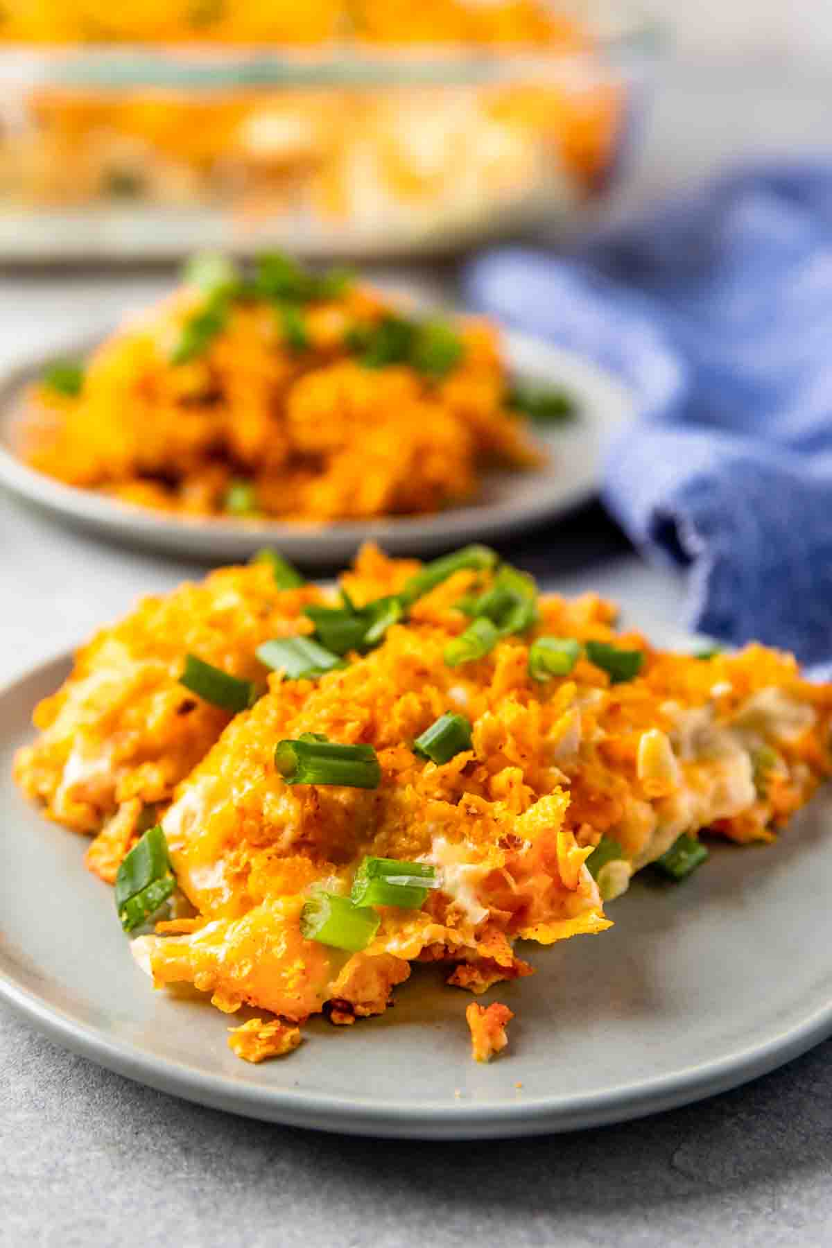 A close-up of a cheesy casserole topped with crushed crackers and chopped green onions on a gray plate, with another serving and a blue napkin in the background.