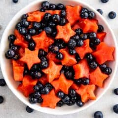 A white bowl filled with star-shaped watermelon pieces and whole blueberries, with extra blueberries scattered on a light surface around the bowl.