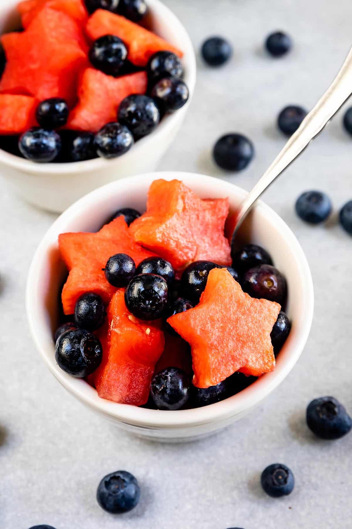 A white bowl filled with blueberries and star-shaped watermelon pieces, with a spoon inside. More blueberries and another bowl with the same fruit are in the background on a light surface.