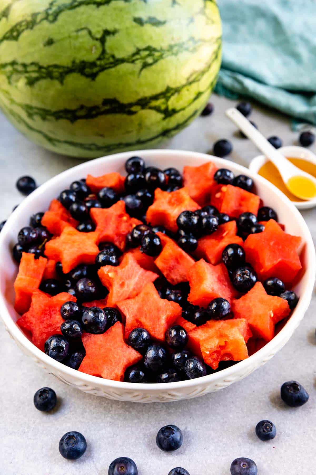A white bowl filled with star-shaped watermelon pieces and fresh blueberries. A whole watermelon, scattered blueberries, a green cloth, and a small dish of honey with a spoon are in the background.