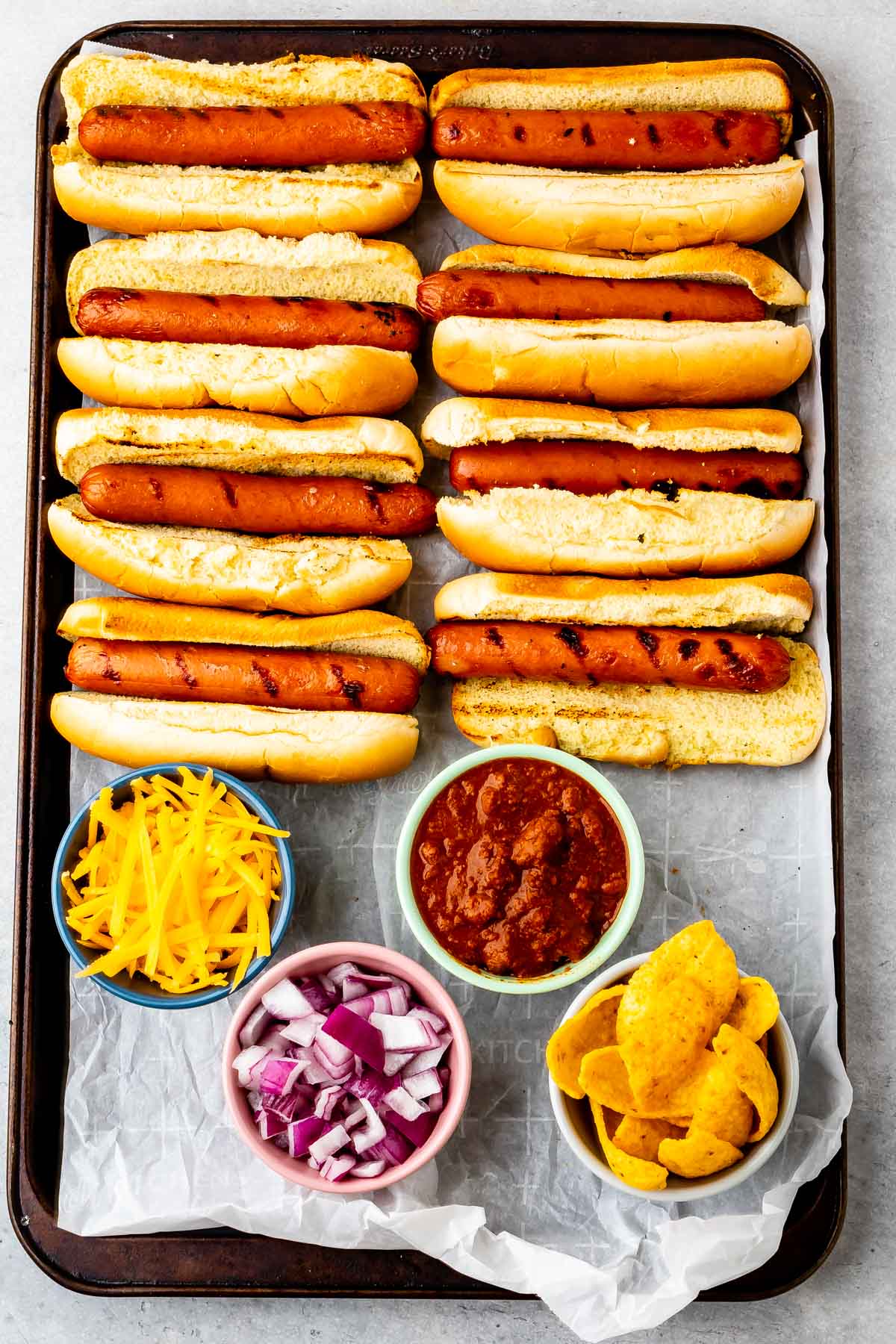 A baking sheet with eight grilled hot dogs in buns, next to small bowls of shredded cheddar cheese, chopped red onions, chili, and corn chips on parchment paper.