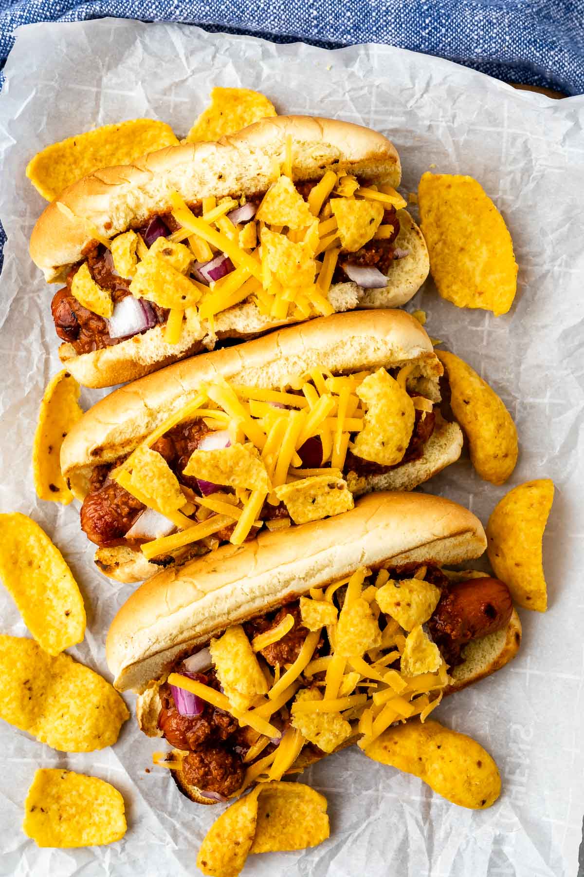 Three chili cheese hot dogs topped with shredded cheddar cheese, corn chips, and chopped onions are arranged on crumpled parchment paper, with extra corn chips scattered around.