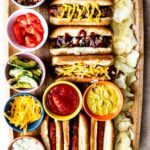 A wooden tray with grilled hot dogs in buns, topped with ketchup, mustard, and cheese. Surrounding bowls contain bacon bits, diced tomatoes, avocado slices, shredded cheese, onions, ketchup, and mustard, with potato chips on the side.