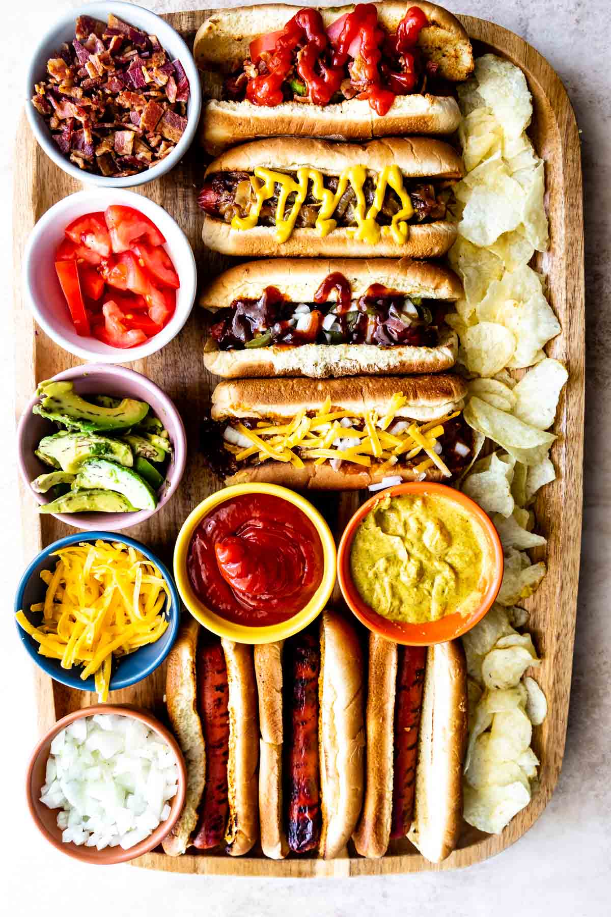 A wooden tray with grilled hot dogs in buns, topped with ketchup, mustard, and cheese. Surrounding bowls contain bacon bits, diced tomatoes, avocado slices, shredded cheese, onions, ketchup, and mustard, with potato chips on the side.