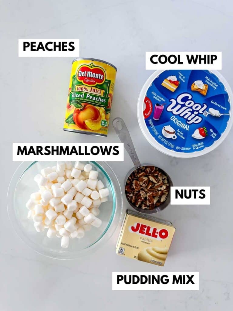 A can of sliced peaches, a tub of Cool Whip, a bowl of mini marshmallows, a measuring cup of chopped nuts, and a box of vanilla pudding mix are arranged on a white surface with labels for each ingredient.