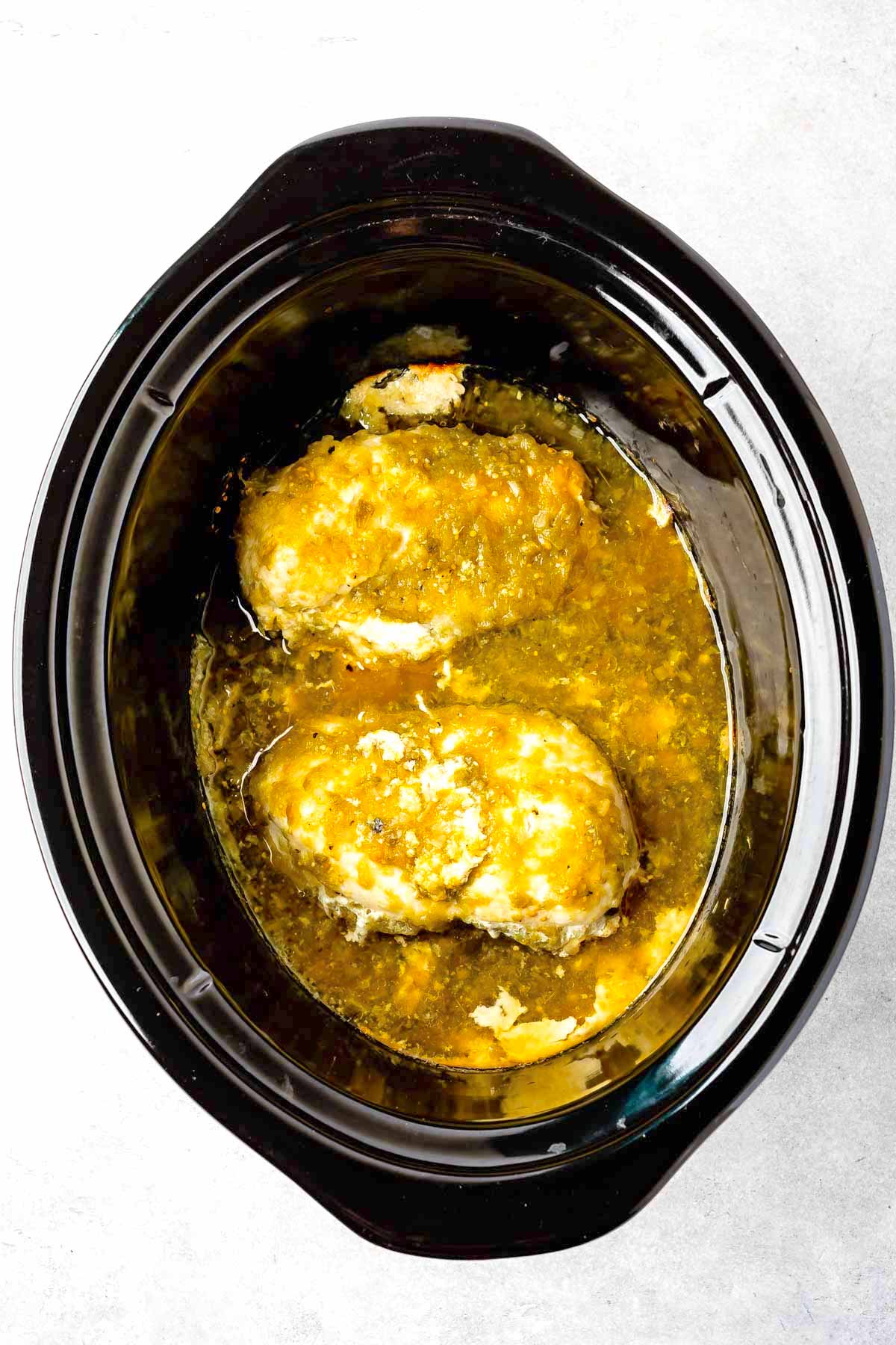 Two pieces of chicken breast covered in a yellow sauce are cooking inside a black slow cooker on a light gray surface.
