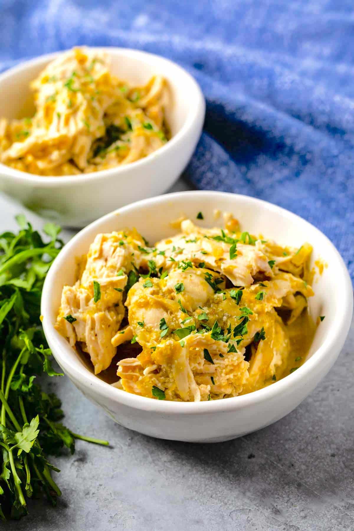 Two white bowls filled with creamy shredded chicken, garnished with chopped parsley, sit on a gray surface with a bunch of fresh parsley nearby and a blue cloth in the background.