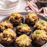 A plate of stuffed mushrooms topped with breadcrumbs and chopped herbs sits on a table, with stacked plates and a striped towel in the background.