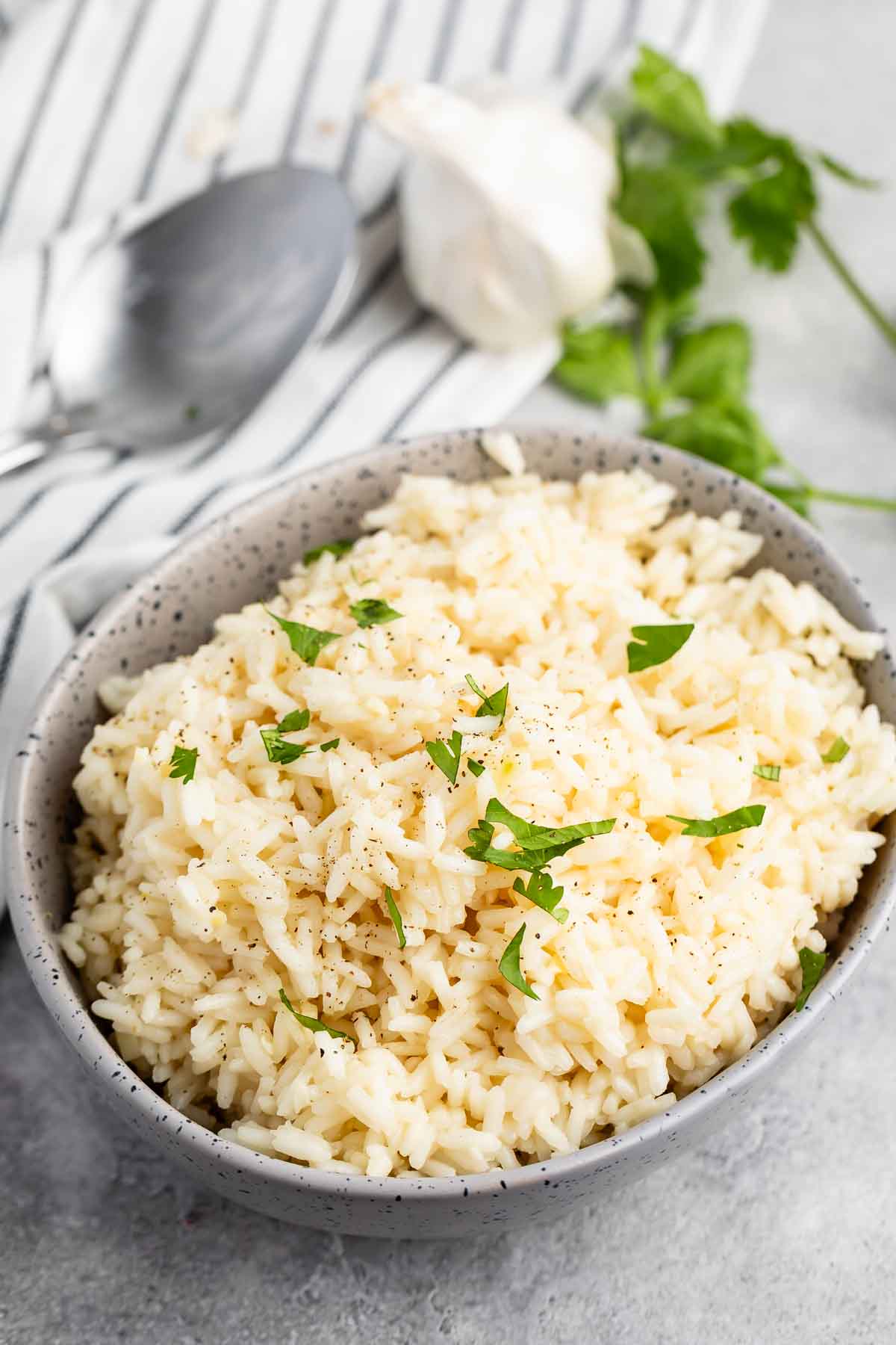 A speckled bowl filled with cooked white rice, garnished with chopped parsley and black pepper, sits on a gray surface. In the background are a spoon, garlic bulb, and parsley leaves on a striped cloth.