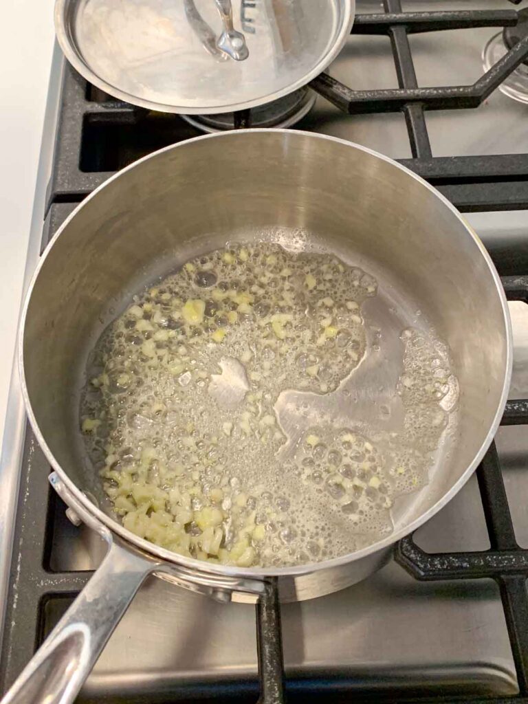 A stainless steel saucepan with minced garlic sautéing in melted butter on a gas stove; a pot lid rests nearby.