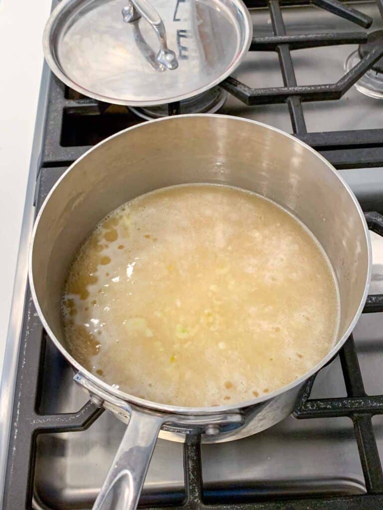 A stainless steel pot on a stove contains a simmering mixture of grains and liquid, with small bubbles and pieces visible. A pot lid rests on the stove nearby.