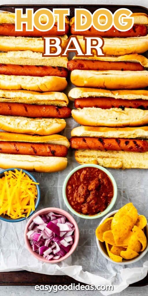 A tray with several grilled hot dogs in buns, surrounded by small bowls of shredded cheese, chopped red onion, chili, and corn chips. The image is labeled HOT DOG BAR at the top.