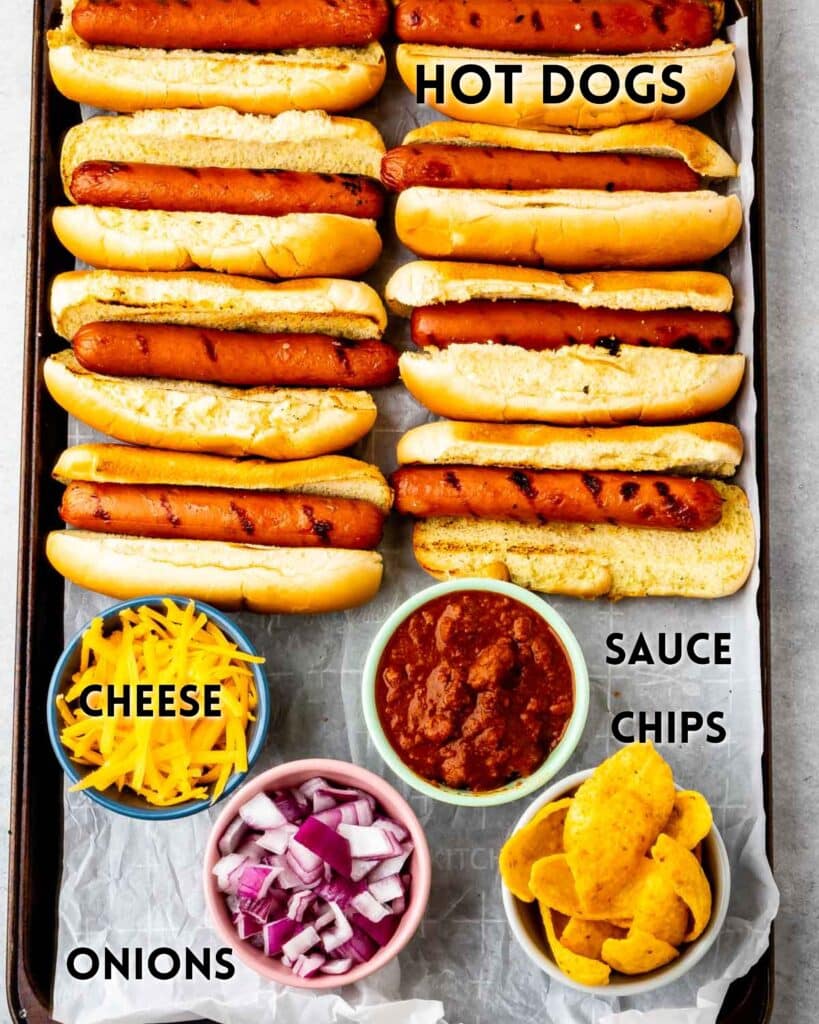 A tray with six hot dogs in buns, and small bowls of shredded cheese, chopped red onions, chili sauce, and corn chips labeled HOT DOGS, CHEESE, ONIONS, SAUCE, and CHIPS.