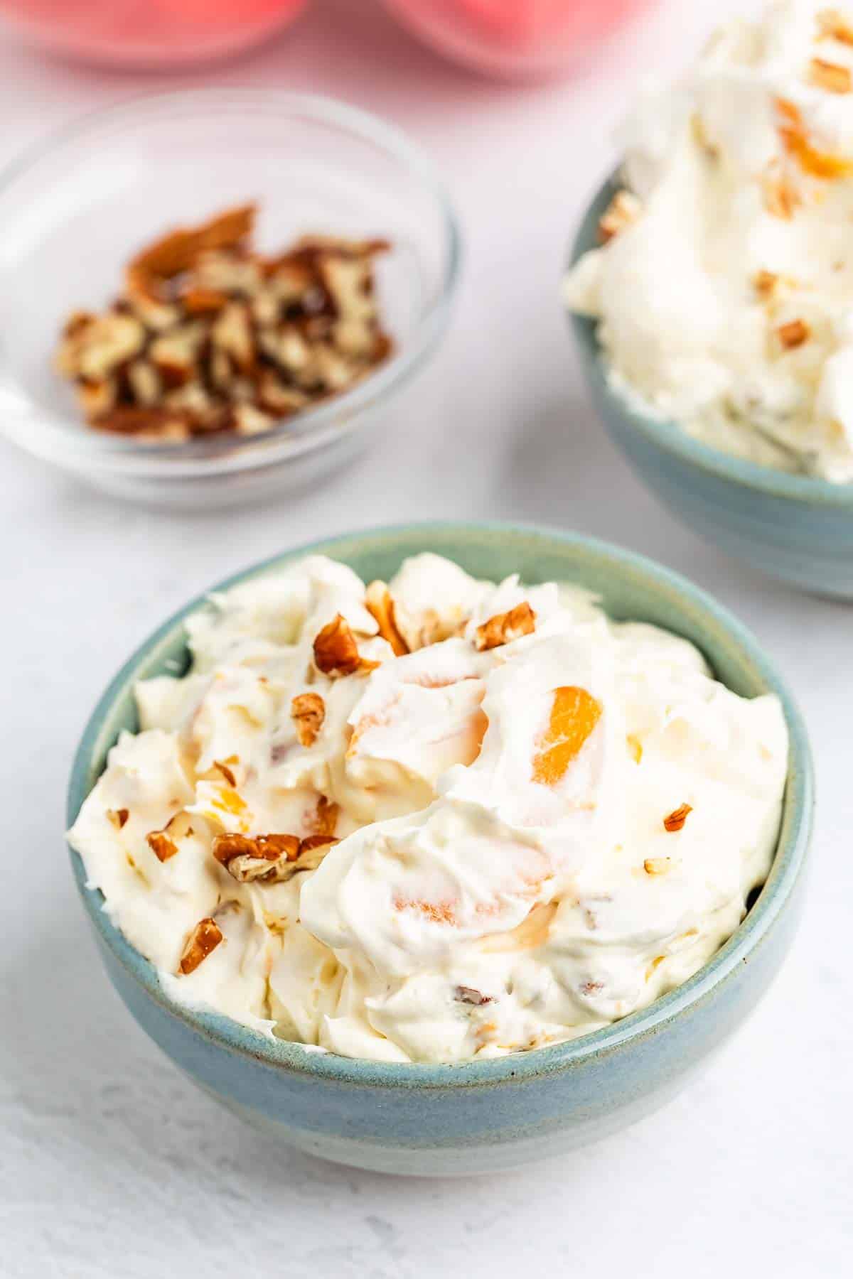 A light blue bowl filled with creamy fruit salad topped with chopped nuts, with another bowl and a small dish of additional nuts in the background on a white surface.