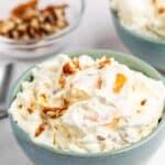 A bowl of creamy fruit salad with whipped topping, orange slices, and chopped nuts, with another bowl and a small dish of nuts in the background.