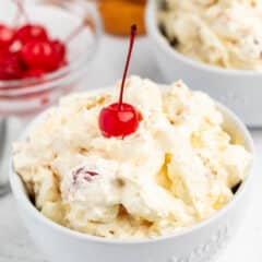A white bowl filled with creamy dessert topped with a red maraschino cherry, with another bowl and a small dish of cherries and cookies in the background.