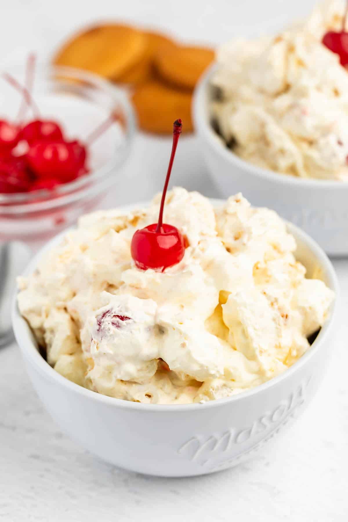 A white bowl filled with creamy dessert topped with a red maraschino cherry, with another bowl and a small dish of cherries and cookies in the background.