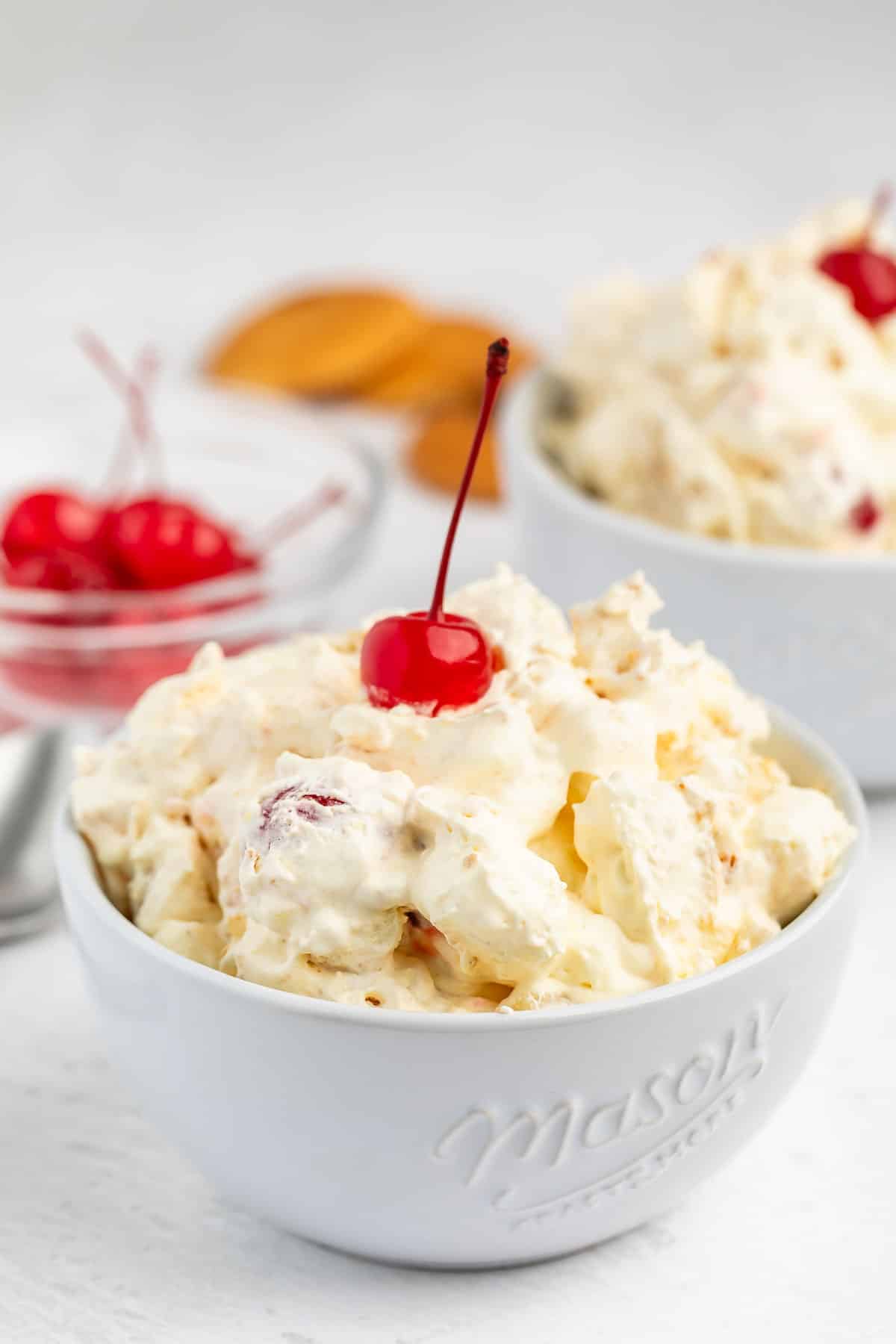 A white bowl filled with creamy ambrosia salad topped with a maraschino cherry. Another bowl and a dish of cherries with cookies are blurred in the background.
