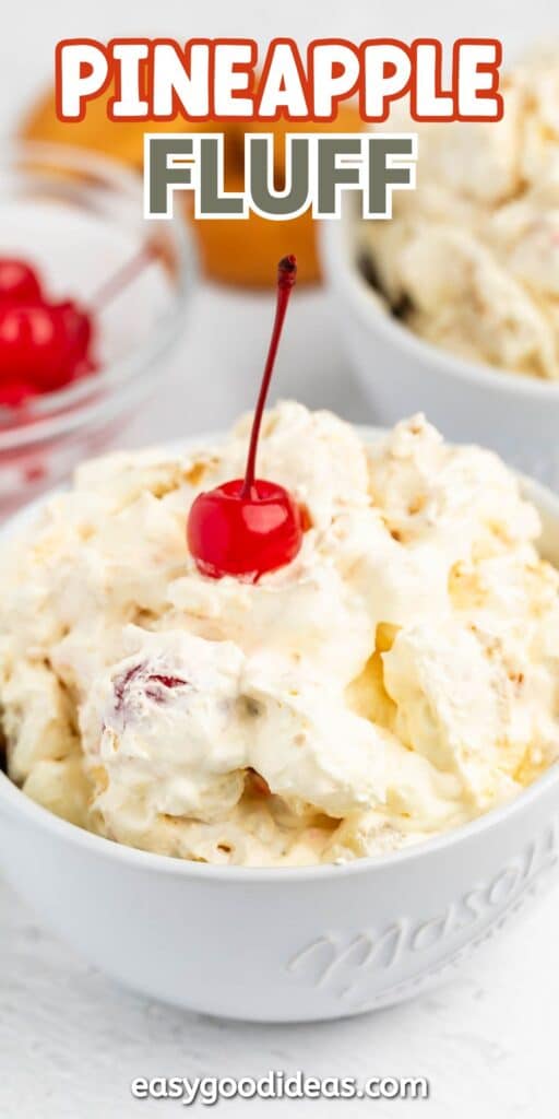 A white bowl filled with creamy pineapple fluff dessert, topped with a red maraschino cherry. Another bowl and a dish of cherries are blurred in the background.