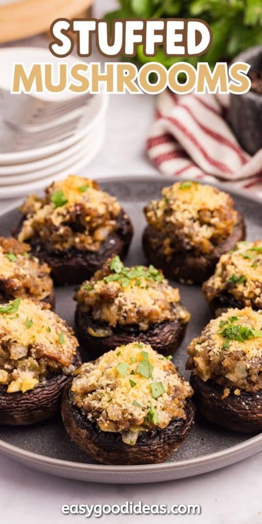 A plate of stuffed mushrooms topped with breadcrumbs and chopped herbs sits on a table, with stacked plates and a striped towel in the background. Text reads Stuffed Mushrooms and easygoodideas.com at the bottom.