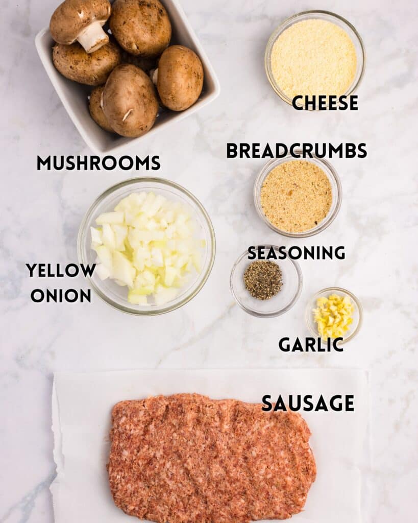 Flat lay of ingredients on a marble surface, labeled: whole mushrooms, shredded cheese, breadcrumbs, diced yellow onion, seasoning, minced garlic, and a slab of ground sausage.