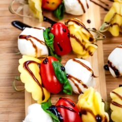 Skewers with mozzarella balls, tortellini, cherry tomatoes, and basil leaves, drizzled with balsamic glaze, are arranged on a wooden board.
