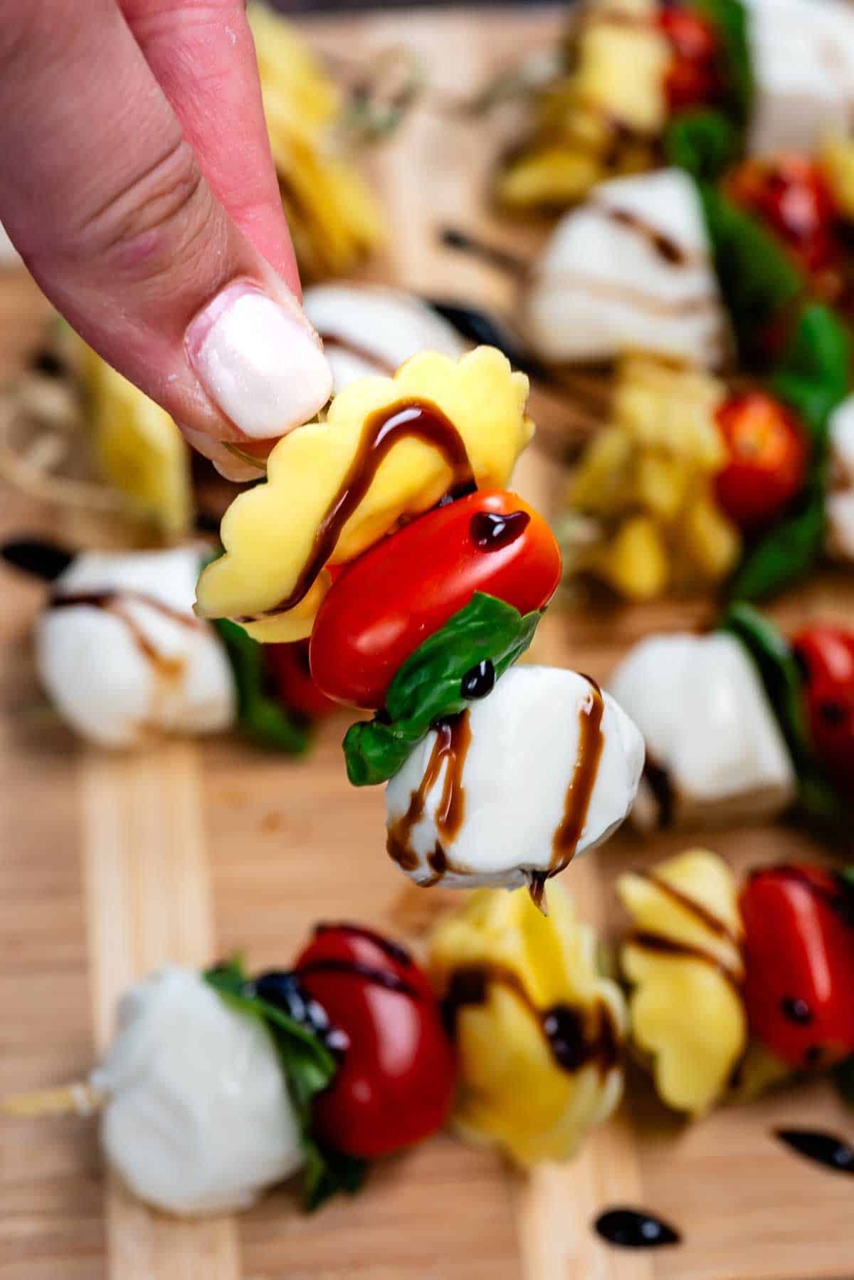 A hand holds a skewer with a cheese-filled ravioli, cherry tomato, fresh basil, and mozzarella ball, all drizzled with balsamic glaze. More skewers are arranged on a wooden board in the background.