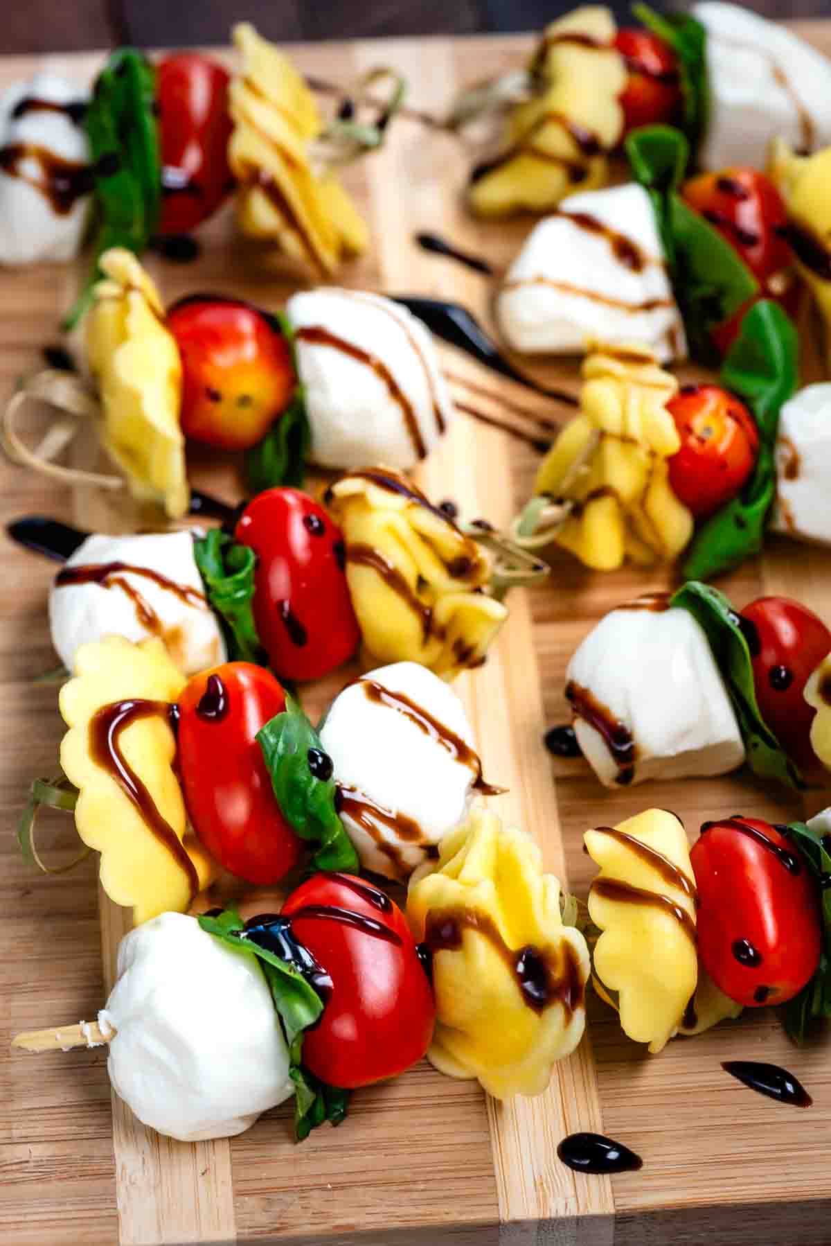 Colorful skewers with yellow tortellini, cherry tomatoes, fresh mozzarella balls, and basil leaves, drizzled with balsamic glaze, arranged on a wooden board.