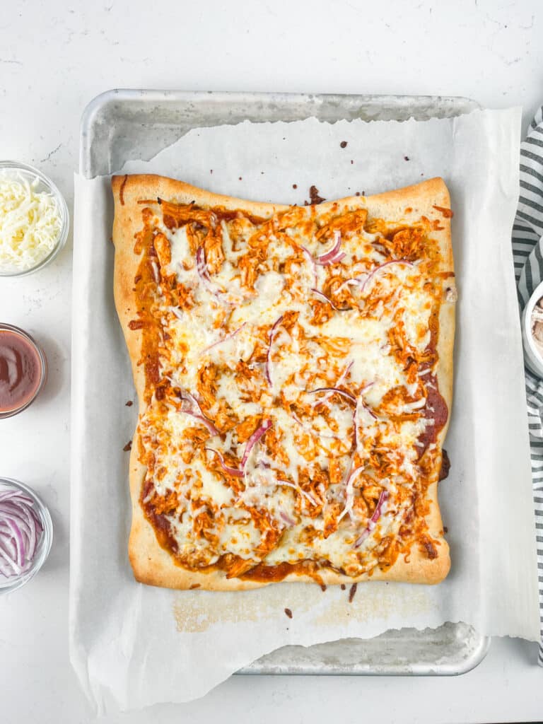 A rectangular homemade pizza with melted cheese, red onions, and barbecue sauce sits on a parchment-lined baking sheet. Small bowls with shredded cheese, barbecue sauce, and sliced onions are nearby on a white surface.