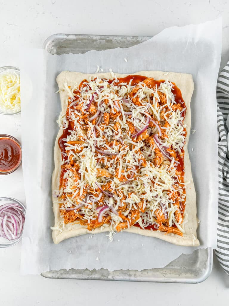 Rectangular pizza dough on a parchment-lined baking sheet topped with tomato sauce, shredded cheese, sliced red onions, and chicken. Small bowls of sauce, onions, and cheese are nearby. A striped towel is at the side.