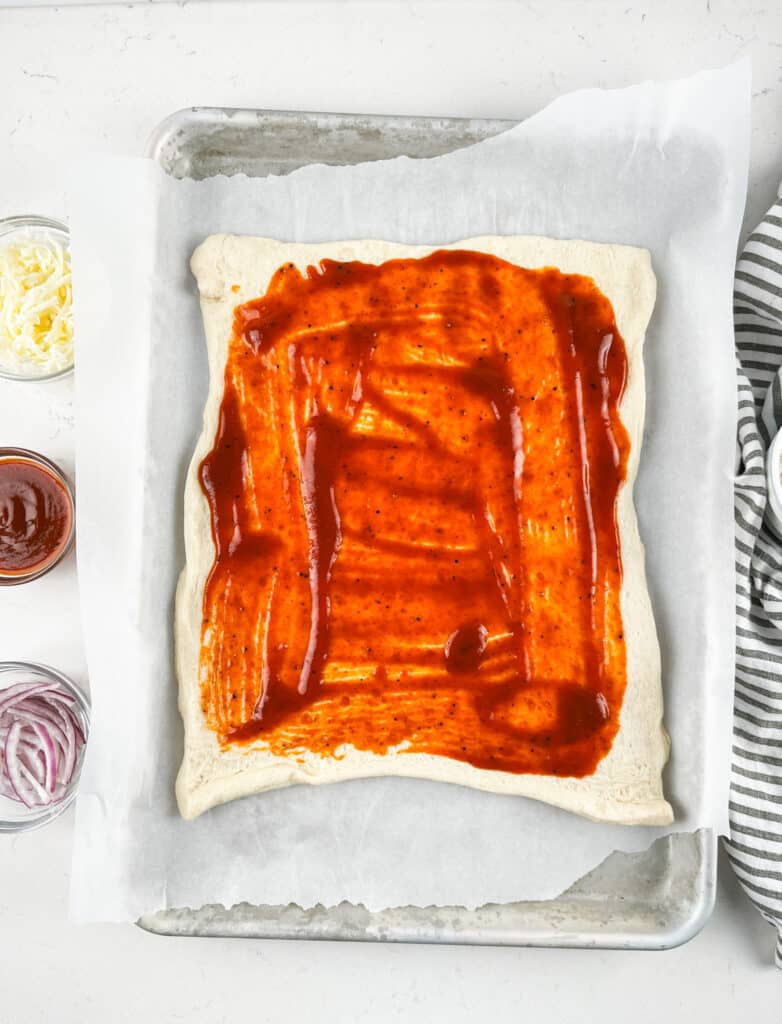 A rectangular sheet of pizza dough on parchment paper is spread with pizza sauce. Nearby are small bowls of shredded cheese, sliced red onions, and more sauce. A striped towel is partially visible on the right.